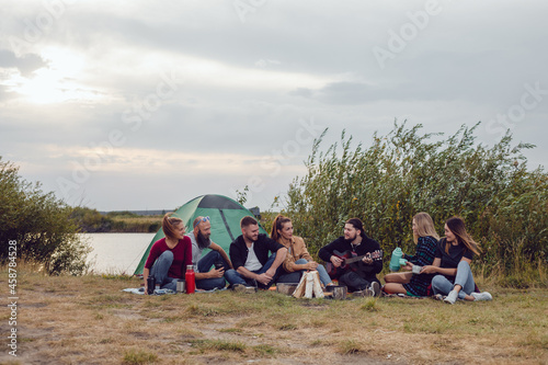 A group of friends sit by the fire on the background of the river and sing to the guitar.