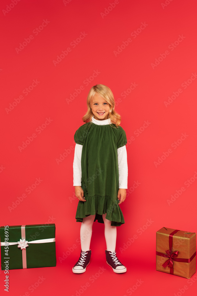 full length view of happy girl in green dress and gumshoes near gift boxes on red