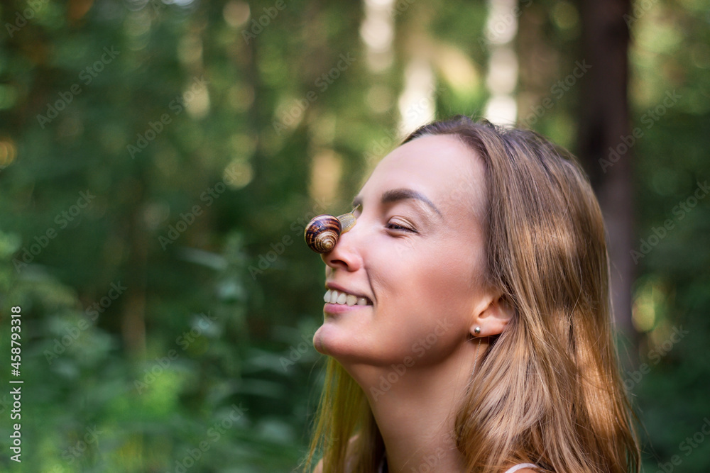 A snail sits on the nose of a Caucasian woman. The girl laughs and looks at the clam. Close-up portrait against the background of a green forest A snail sits on the nose of a Caucasian woman. The girl laughs and looks at the clam. Close-up portrait against the background of a green forest