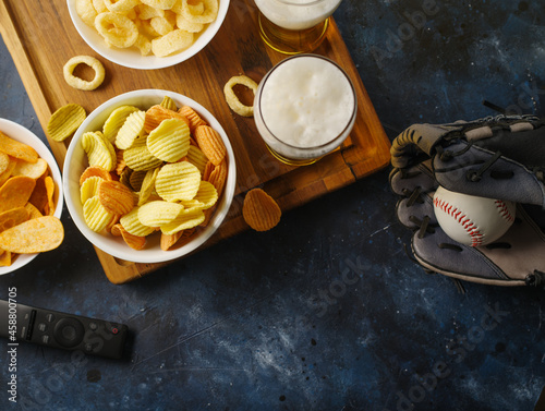 A classic set of sports fans - on a wooden tray, glasses of beer, snacks, potato chips, onion rings. Nearby lies a leather baseball glove and a ball. Watching sports games on TV with friends.