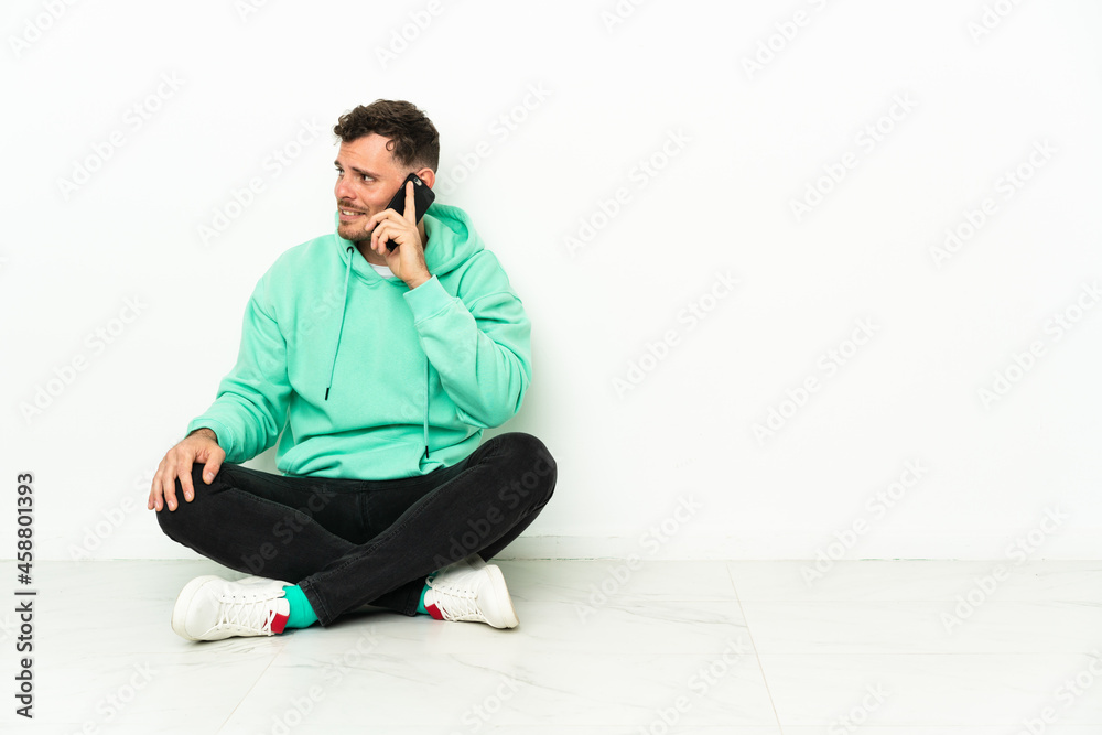 Young handsome caucasian man sitting on the floor keeping a conversation with the mobile phone with someone Young handsome caucasian man sitting on the floor keeping a conversation with the mobile phone with someone