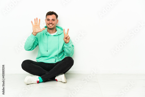 Young handsome caucasian man sitting on the floor counting seven with fingers