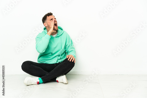 Young handsome caucasian man sitting on the floor shouting with mouth wide open to the lateral