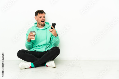 Young handsome caucasian man sitting on the floor holding coffee to take away and a mobile