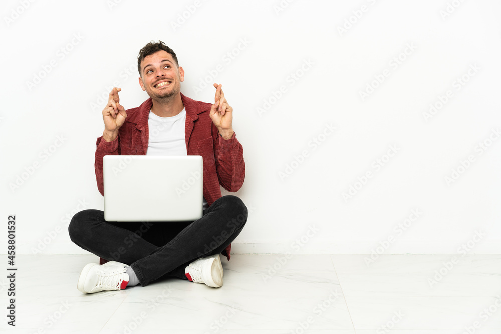 Young handsome caucasian man sit-in on the floor with laptop with fingers crossing Young handsome caucasian man sit-in on the floor with laptop with fingers crossing