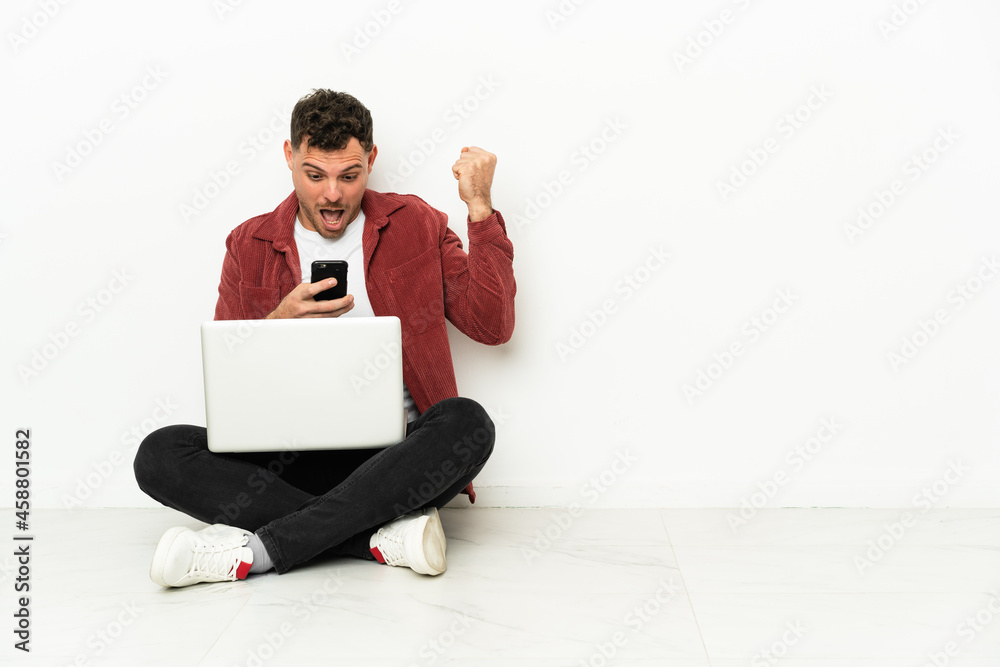 Young handsome caucasian man sit-in on the floor with laptop surprised and sending a message Young handsome caucasian man sit-in on the floor with laptop surprised and sending a message