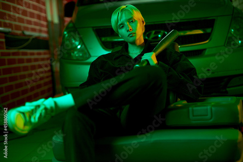 a young girl with a bat sits at the hood of a car and looks straight into the camera. Modern stylish shooting in neon light. High quality photo
