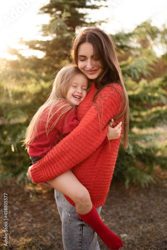 Happy woman hold little kid girl 3-4 year old wear red bright sweaters posing over nature background close up. Family lifestyle concept. Motherhood. Autumn season. Daughter and mother together.
