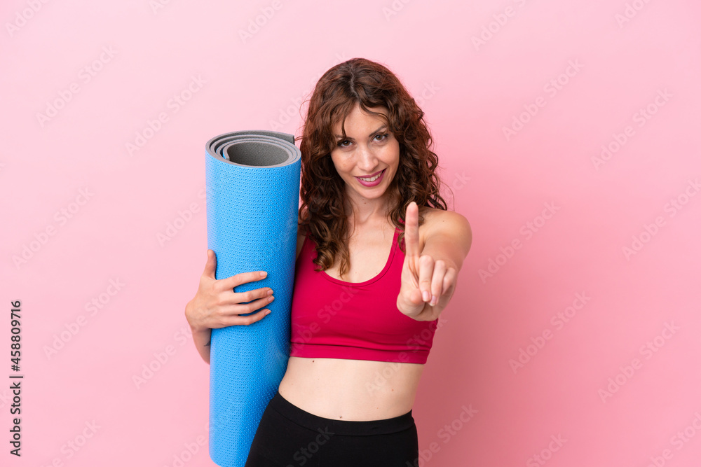 Young sport woman going to yoga classes while holding a mat isolated on pink background showing and lifting a finger Young sport woman going to yoga classes while holding a mat isolated on pink background showing and lifting a finger