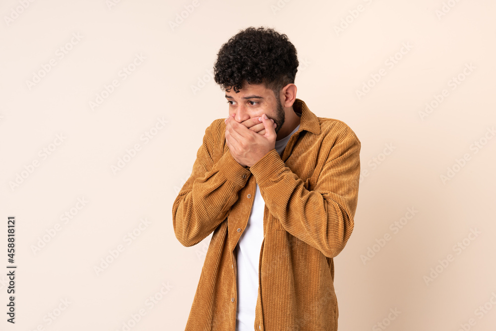 Young Moroccan man isolated on beige background covering mouth and looking to the side Young Moroccan man isolated on beige background covering mouth and looking to the side