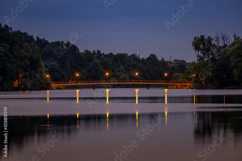 Fototapeta Naklejka Na Ścianę i Meble -  The bridge on the Dlugie Lake is illuminated by the light from the lamps after sunset. The light from the lamps is reflected in the water and leaves long yellow streaks. - Olsztyn, Warmia and Mazury, 