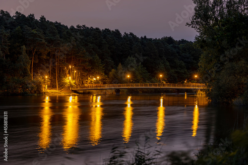 The bridge on the Dlugie Lake is illuminated by the light from the lamps after sunset. The light from the lamps is reflected in the water and leaves long yellow streaks. - Olsztyn, Warmia and Mazury,  © Grzej