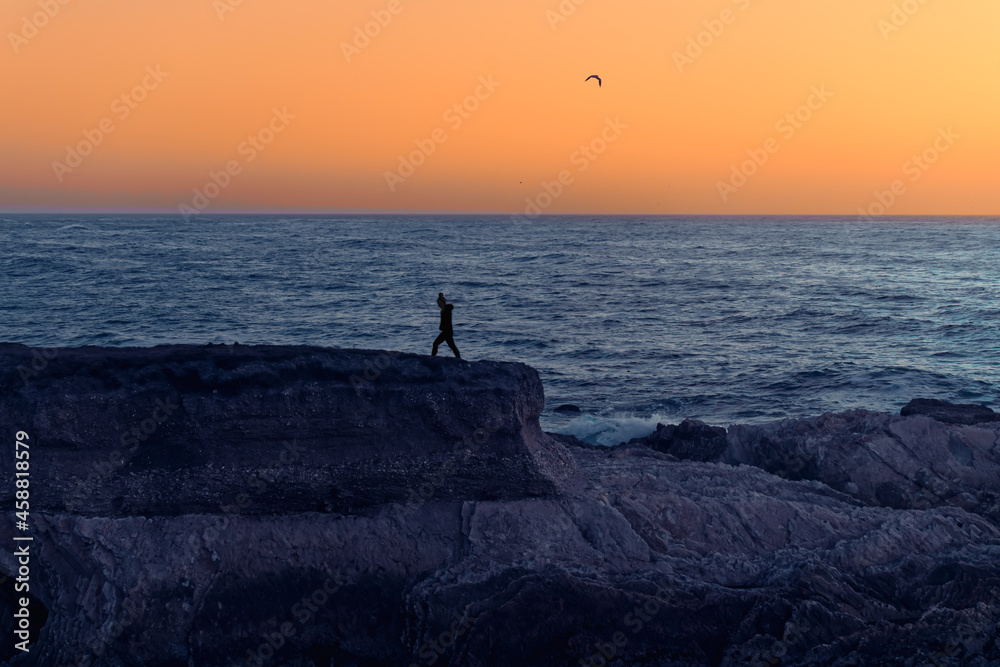 Sunset seascape. Rocky cliffs and silhouette of walking woman, blue ocean and bright yellow sky on background
