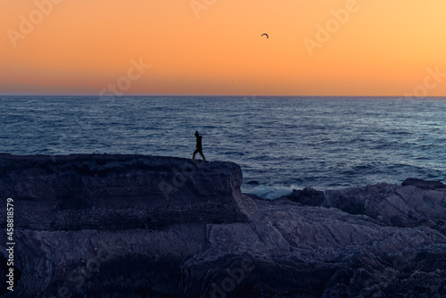 Sunset seascape. Rocky cliffs and silhouette of walking woman, blue ocean and bright yellow sky on background