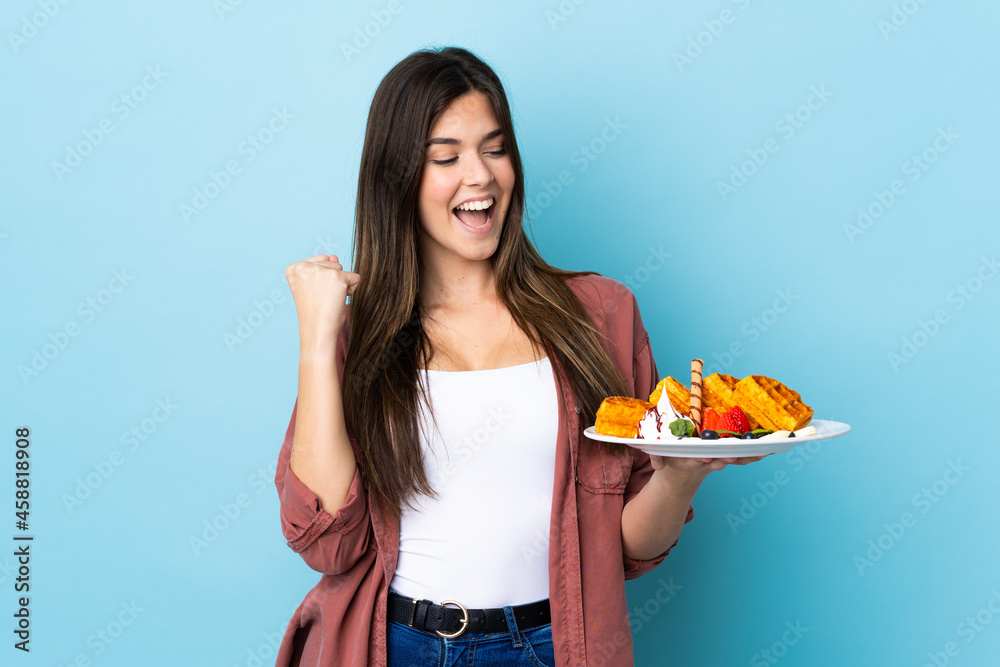 Teenager Brazilian girl holding waffles over isolated blue background celebrating a victory Teenager Brazilian girl holding waffles over isolated blue background celebrating a victory