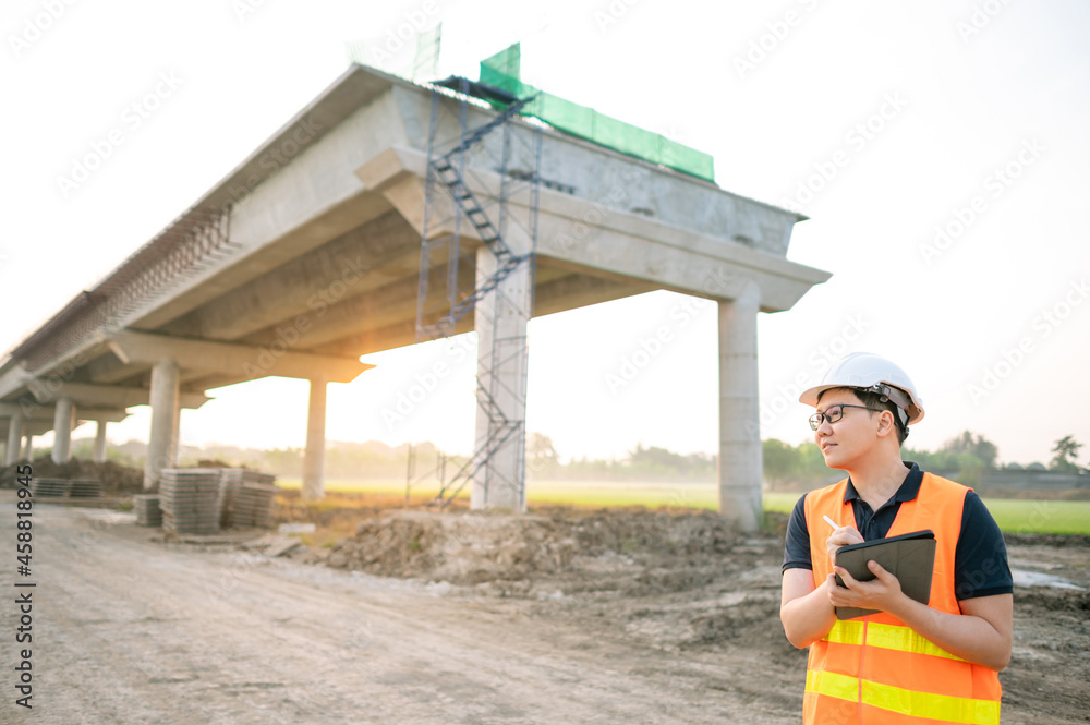 Smart Asian worker man or male civil engineer with protective safety helmet and reflective vest using digital tablet for project planning and checking architectural drawing at construction site. Smart Asian worker man or male civil engineer with protective safety helmet and reflective vest using digital tablet for project planning and checking architectural drawing at construction site.