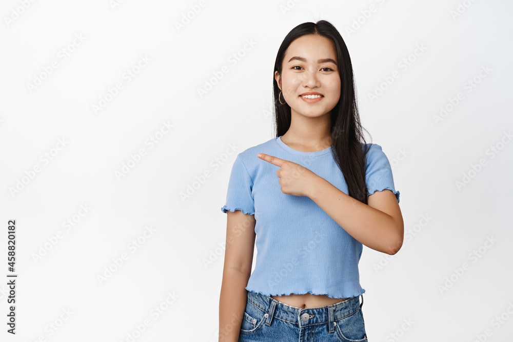 Cute asian girl pointing left and smiling, showing direction, sale advertisement, standing in blue tshirt over white background Cute asian girl pointing left and smiling, showing direction, sale advertisement, standing in blue tshirt over white background