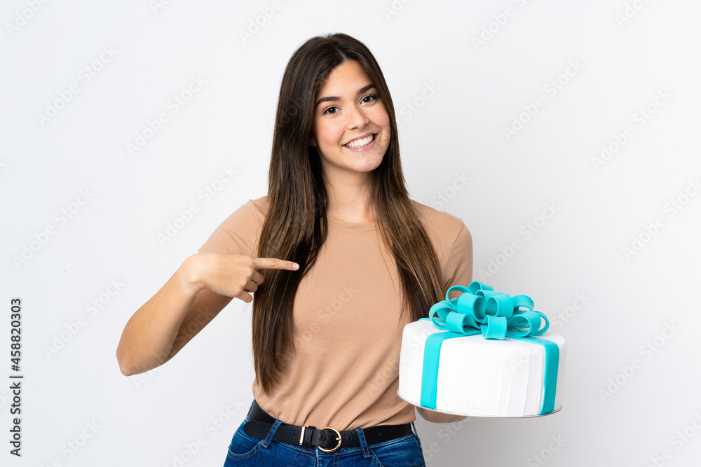 Teenager pastry chef holding a big cake over isolated white background and pointing it Teenager pastry chef holding a big cake over isolated white background and pointing it