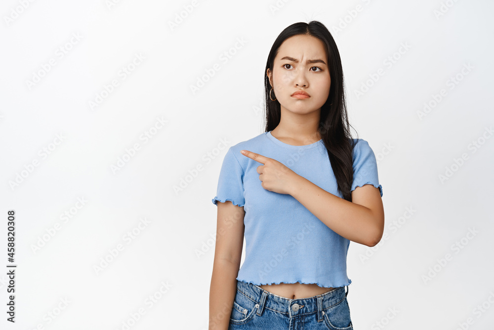 Angry asian girl pointing, looking left with grumpy frowning face, stare with dislike, standing in tshirt over white background Angry asian girl pointing, looking left with grumpy frowning face, stare with dislike, standing in tshirt over white background