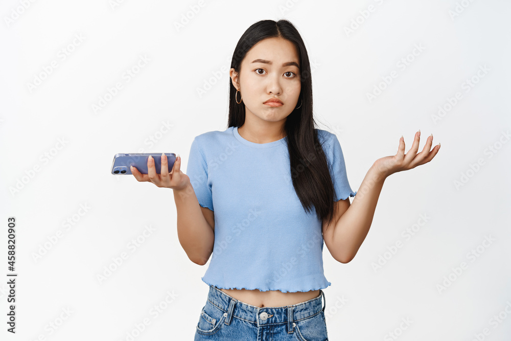 Confused asian girl holding smartphone and shrugging shoulders indecisive, dont know, cant understand, standing over white background Confused asian girl holding smartphone and shrugging shoulders indecisive, dont know, cant understand, standing over white background