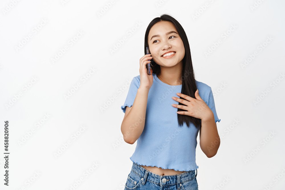 Happy smiling asian girl answer phone call, talking on cellphone, standing over white background Happy smiling asian girl answer phone call, talking on cellphone, standing over white background