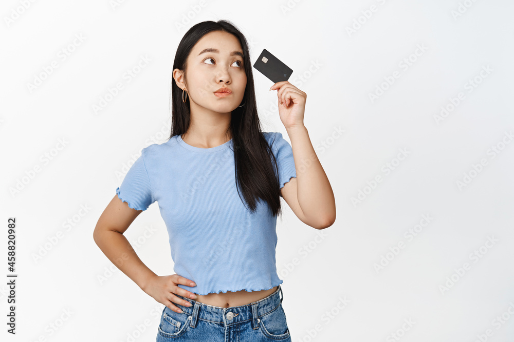 Silly and cute asian girl looking at credit card and thinking, standing thoughtful against white background Silly and cute asian girl looking at credit card and thinking, standing thoughtful against white background