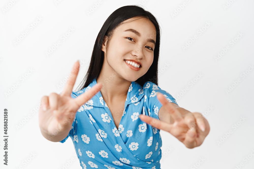 Beautiful asian female model showing peace v-sign gestures and smiling at camera, wearing blue dress, standing over white background Beautiful asian female model showing peace v-sign gestures and smiling at camera, wearing blue dress, standing over white background
