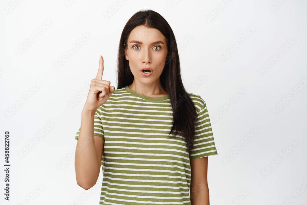 Surprised brunette woman staring impressed at camera, pointing finger up, amazed by something upwards, standing over white background Surprised brunette woman staring impressed at camera, pointing finger up, amazed by something upwards, standing over white background