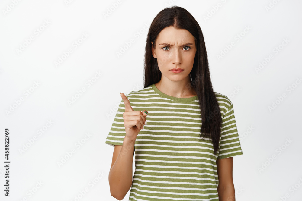Angry brunette woman shaking finger in disapproval, scolding bad behaviour, standing over white background in t-shirt