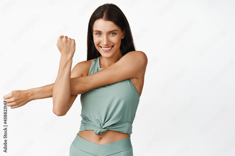Happy fitness woman stretch her arms and smiling, warm up before workout and jogging training session, standing in activewear against white background Happy fitness woman stretch her arms and smiling, warm up before workout and jogging training session, standing in activewear against white background