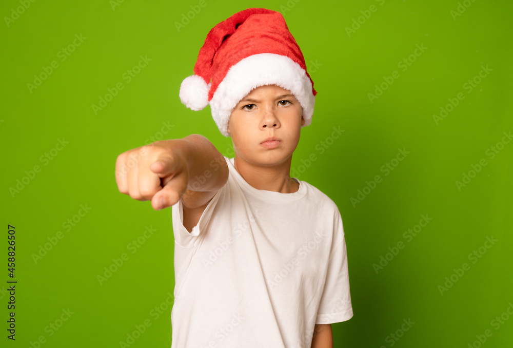 Portrait of a serious boy in a red hat and white t-shirt pointing finger to camera on a green background. A child in a Christmas hat of Santa Claus. Portrait of a serious boy in a red hat and white t-shirt pointing finger to camera on a green background. A child in a Christmas hat of Santa Claus.