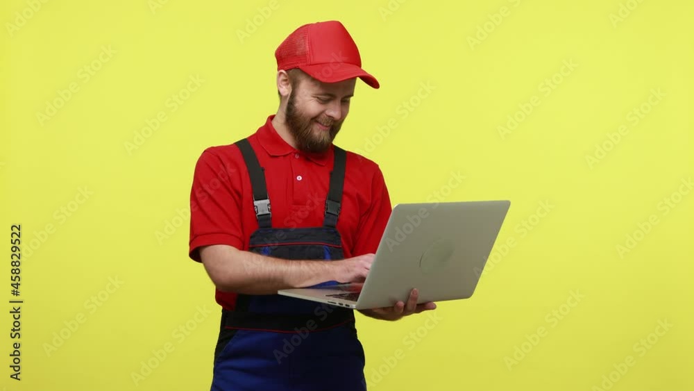 Positive technician working with laptop computer, looking at display, expressing positive emotions, wearing blue uniform, red T-shirt and visor cap. Indoor studio shot isolated over yellow background. Positive technician working with laptop computer, looking at display, expressing positive emotions, wearing blue uniform, red T-shirt and visor cap. Indoor studio shot isolated over yellow background.