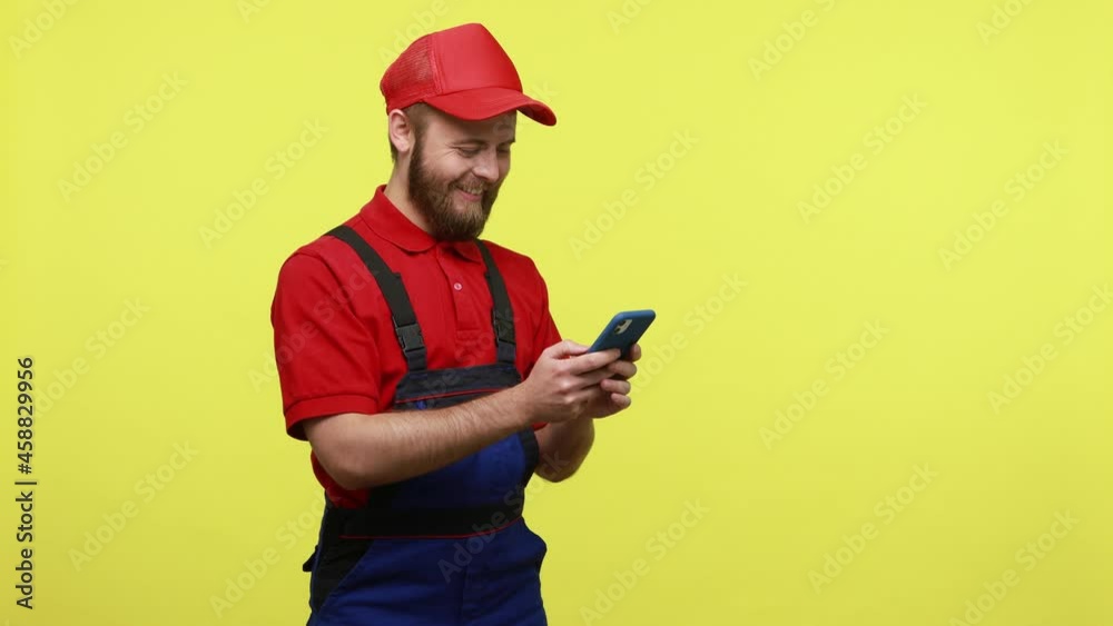 Side view of happy smiling workman using mobile phone and laughing, typing message for his client, wearing blue uniform, red T-shirt and visor cap. Indoor studio shot isolated over yellow background. Side view of happy smiling workman using mobile phone and laughing, typing message for his client, wearing blue uniform, red T-shirt and visor cap. Indoor studio shot isolated over yellow background.