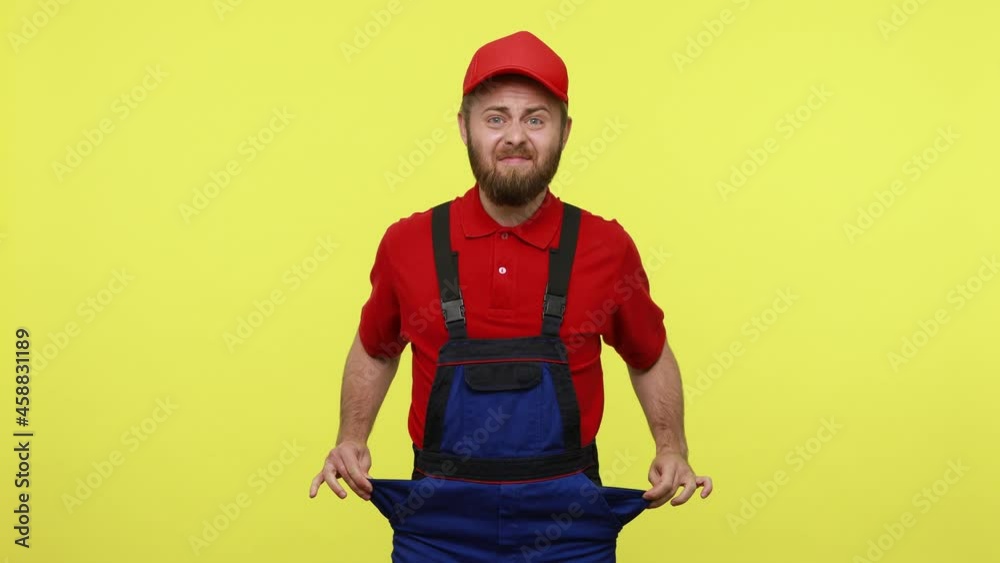 Handsome workman showing his empty porckets, expressing sadness, has no orders and salary, wearing blue uniform, red T-shirt and visor cap. Indoor studio shot isolated over yellow background. Handsome workman showing his empty porckets, expressing sadness, has no orders and salary, wearing blue uniform, red T-shirt and visor cap. Indoor studio shot isolated over yellow background.