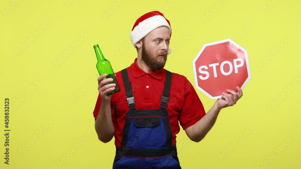 Handsome bearded workman holding bottle of alcohol and red stop sign, do not drink at work, wearing blue overalls and santa claus hat. Indoor studio shot isolated over yellow background. Handsome bearded workman holding bottle of alcohol and red stop sign, do not drink at work, wearing blue overalls and santa claus hat. Indoor studio shot isolated over yellow background.