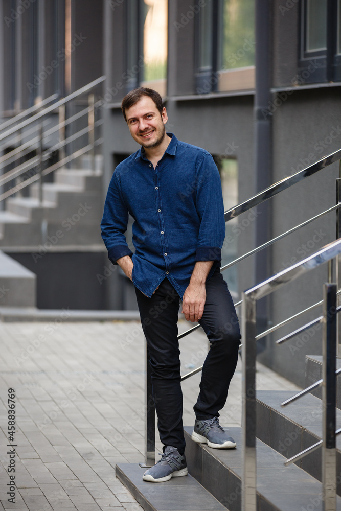 Young man weared in blue denim shirt on background of the City. Fashion style guy classic portrait. Street style male portrait looking to camera. Contemporary businessman. Modern urban walk in a town Young man weared in blue denim shirt on background of the City. Fashion style guy classic portrait. Street style male portrait looking to camera. Contemporary businessman. Modern urban walk in a town