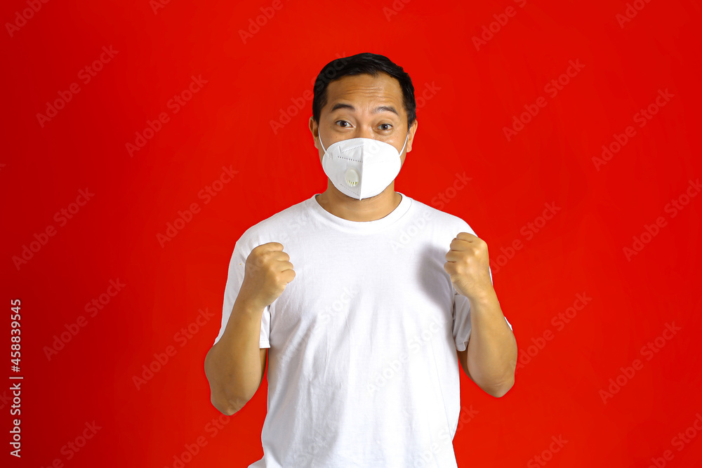 Asian man wearing a medical mask, clenched his hands while looking at the camera with an expression of success on a red background Asian man wearing a medical mask, clenched his hands while looking at the camera with an expression of success on a red background