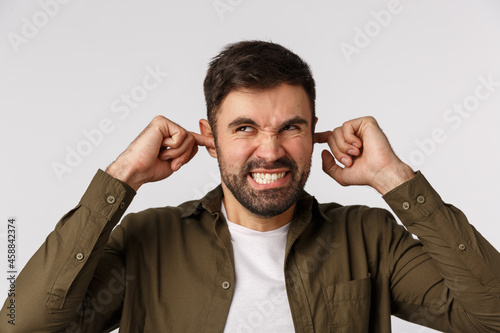 Cant stand this annoying stupid neigbours with theis sound system. Angry and irritated, annoyed bearded guy cursing and grimacing annoyed, closed ears with fingers stare up, disturbed loud music photo