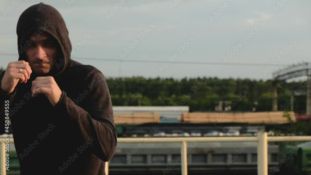 Young focused male athlete in hood aggressively looks at camera and boxes with his hands. Athlete training on roof in evening at sunset. Hard cardio exercises. boxer trains for fight Young focused male athlete in hood aggressively looks at camera and boxes with his hands. Athlete training on roof in evening at sunset. Hard cardio exercises. boxer trains for fight