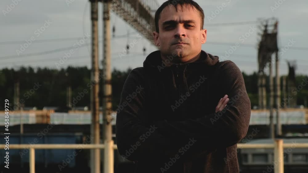 Portrait of serious male athlete on roof in evening at sunset during training. male in hoodie looks intently at camera outside against background of trains with his arms crossed Portrait of serious male athlete on roof in evening at sunset during training. male in hoodie looks intently at camera outside against background of trains with his arms crossed