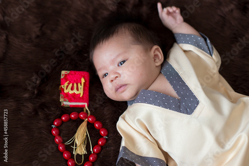 A cute Asian infant Muslim in thobe and rosary beads on red, muslim bead on bed in the home, translation is the name of muslim God 