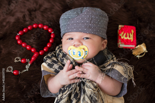 A cute Asian infant Muslim in thobe with prayer cap on bed in home, translation is noble quran