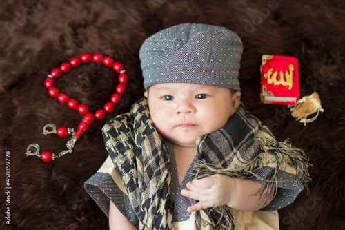 A cute Asian infant Muslim in thobe with prayer cap on bed in home. photo