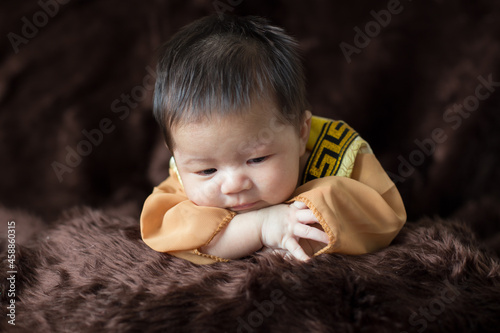 A newborn baby wearing a Chinese emperor's costume gold colour lying on a brown blanket.