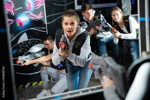 Excited girl aiming laser gun at other players during lasertag game in dark room..