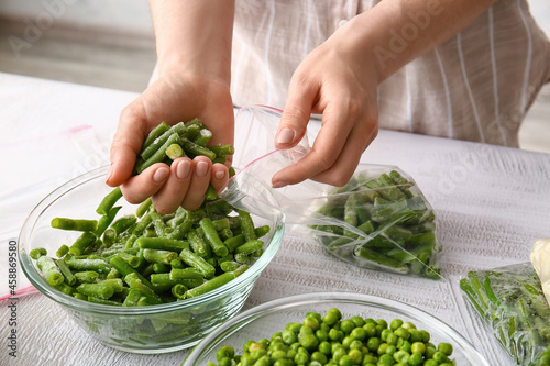 Woman putting frozen green beans in plastic bag at table in kitchen, closeup
