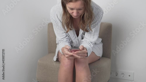 view of pretty brunetter young woman sitting armchair uses phone and smiles at modern room. Texting sharing messages on social media enjoying feel happy mobile technology evening photo