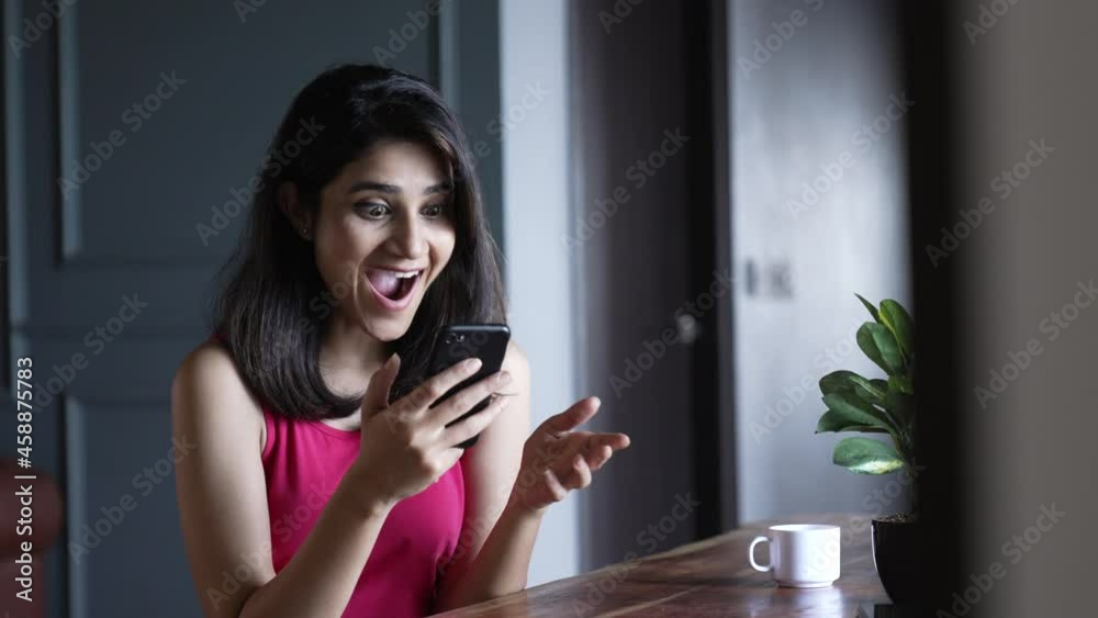 Young woman smiling and laughing while watching a funny video on a smartphone or cellphone.Indian girl holding smart phone watching funny videos,movie or social media content while sitting in home . Young woman smiling and laughing while watching a funny video on a smartphone or cellphone.Indian girl holding smart phone watching funny videos,movie or social media content while sitting in home .