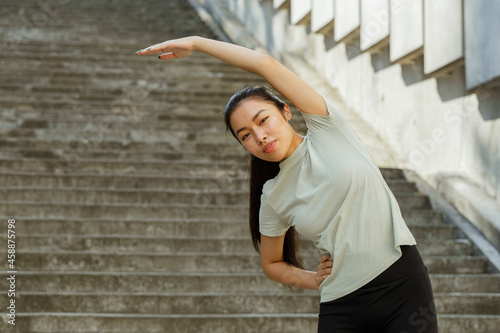 Long haired Asian sportswoman in tracksuit and sneakers does side bend training near wall with concrete decor on city street