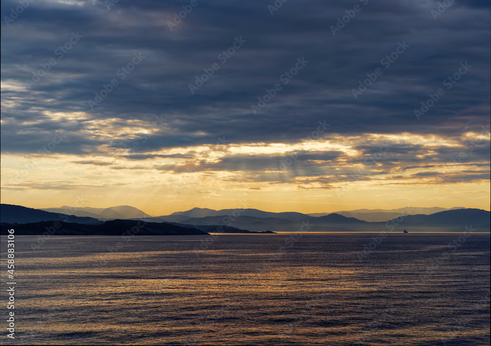 Rays of sunlight penetrating the low cloud layer above the Mountains and Islands of Bergen Fjord, with a Vessel in the Distance heading to Port.